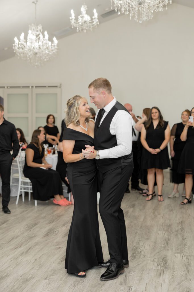 Groom sharing a mother-son dance with his mom beneath chandeliers at Brighton Abbey, captured by Bushel + Peck Photo