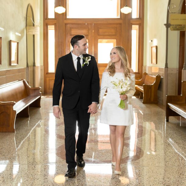 Newlyweds walking hand in hand down a marble hallway inside the Tarrant County Courthouse, captured by Bushel + Peck Photo
