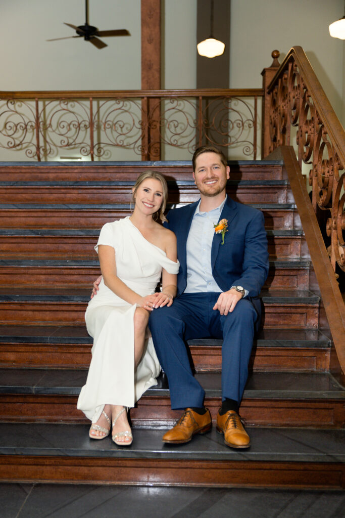 Bride and groom seated together on the carved wooden staircase inside the Tarrant County Courthouse, captured by Bushel + Peck Photo