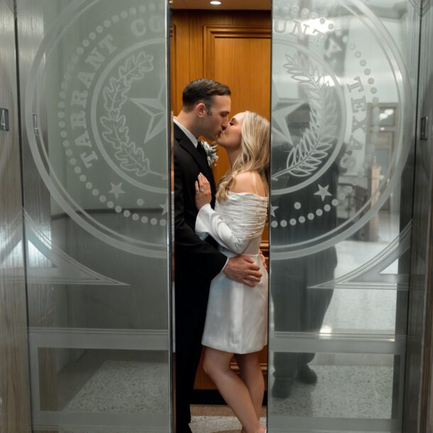 Couple kissing between glass doors etched with the Tarrant County seal inside the courthouse, captured by Bushel + Peck Photo