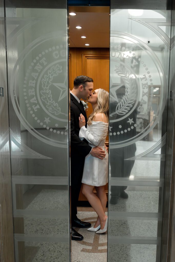 Couple kissing between glass doors etched with the Tarrant County seal inside the courthouse, captured by Bushel + Peck Photo