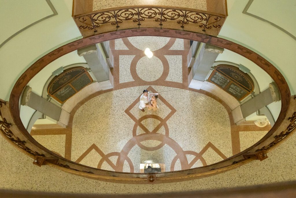 Looking down into the Tarrant County Courthouse rotunda at a couple standing on the terrazzo floor, captured by Bushel + Peck Photo