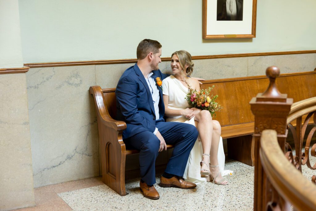 Bride and groom sitting on a wooden bench in a hallway of the Tarrant County Courthouse, captured by Bushel + Peck Photo