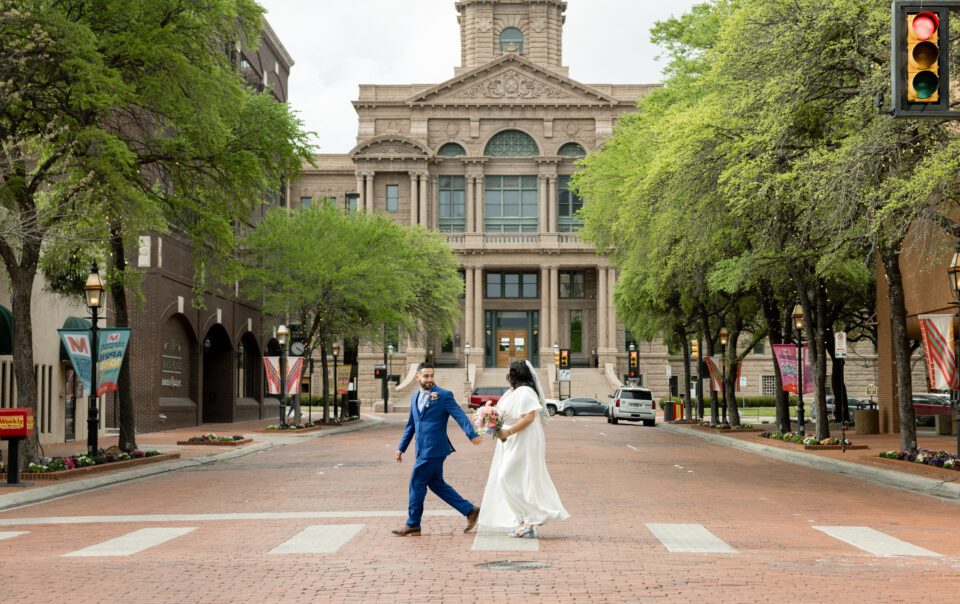 Newlyweds crossing Main Street in downtown Fort Worth with the historic Tarrant County Courthouse behind them, captured by Bushel + Peck Photo
