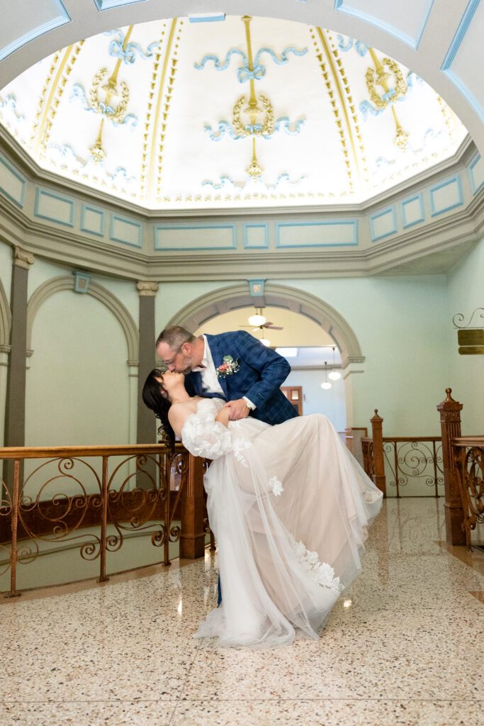 Groom dipping his bride beneath the painted dome of the Tarrant County Courthouse rotunda, captured by Bushel + Peck Photo
