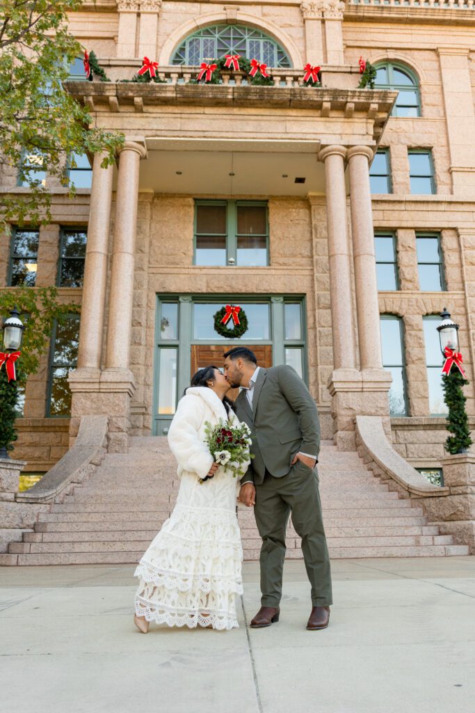 Couple kissing at the Tarrant County Courthouse entrance decorated with holiday wreaths, captured by Bushel + Peck Photo