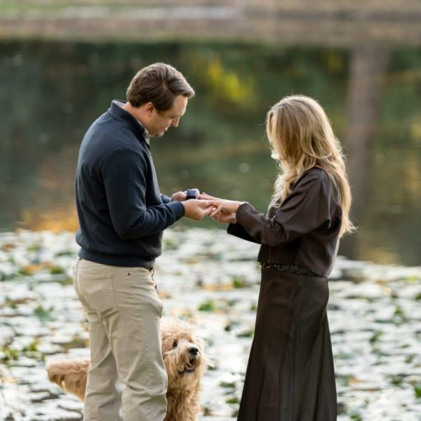 Proposal photo of couple holding hands, exchanging rings at lakeside park. captured by Bushel + Peck Photo