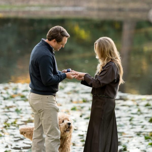 Proposal photo of couple holding hands, exchanging rings at lakeside park. captured by Bushel + Peck Photo