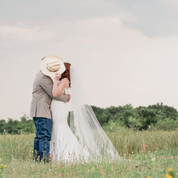 Bride and groom share a romantic kiss in an East Texas field, with the groom’s cowboy hat playfully shielding their faces. The bride’s flowing veil and the rustic landscape highlight this charming East Texas photography location.