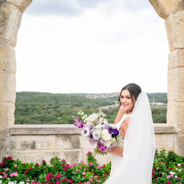 Smiling bride posing with purple and white bouquet at a scenic South Central Texas photography location near San Antonio, Austin, and Houston, framed by stone arches and rolling hills.