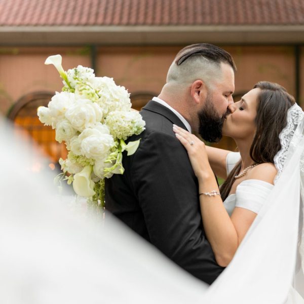 Groom kissing the bride as her veil sweeps across the frame at Rosewood Mansion in Dallas, captured by Bushel + Peck Photo