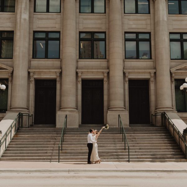 Bride raising her bouquet beside the groom on the steps of a columned courthouse after their micro wedding, captured by Bushel + Peck Photo